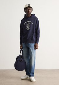 Felpa navy con logo bianco, jeans azzurri chiari, cappello crema con logo e sneaker bianche. Porta una borsa rotonda navy con due manici.
