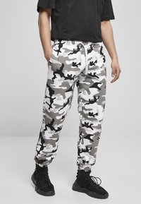 Grau-weiße und schwarze Camouflage-Sweatpants mit elastischem Bund, gerippten Knöcheln und seitlichen Taschen, getragen mit schwarzen Sneakers.