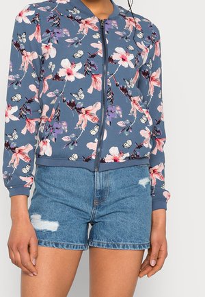 Femme portant une veste zippée bleue à motifs floraux et un short en jean déchiré, debout devant un fond clair uni.