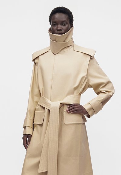 Personne à la peau foncée portant un trench-coat beige ceinturé avec un col montant et des poches à rabat, debout avec une main sur la hanche.