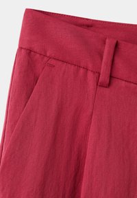 Détail d’un pantalon rouge taille haute montrant la ceinture, la boucle de ceinture, la poche latérale et un pli cousu sur un tissu à texture lisse.