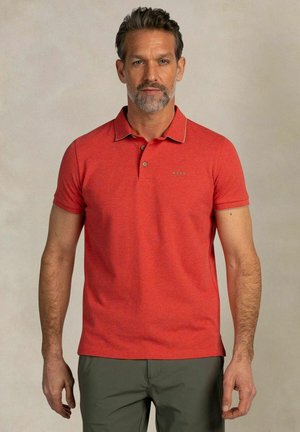 Man met korte baard, een rood poloshirt en olijfgroene broek, staand tegen een neutrale achtergrond.