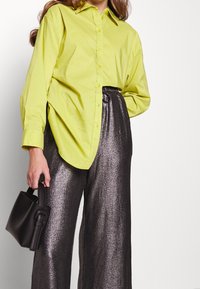 Limegrön knäppskjorta i kombination med metallic silver vida byxor. Modell som håller en liten svart handväska med ett svängt handtag.