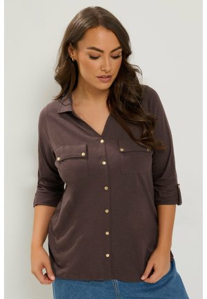 Vrouw met lang bruin haar die een bruine blouse met borstzakken en een blauwe spijkerbroek draagt, kijkt met een neutrale uitdrukking naar beneden.