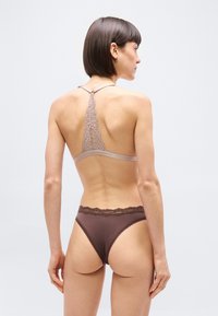 Γυναίκα με κοντά σκούρα μαλλιά, που φαίνεται από πίσω, φοράει μπορντό στρινγκ με δαντέλα και μπεζ δαντελένιο σουτιέν racerback, σε ουδέτερο φόντο.