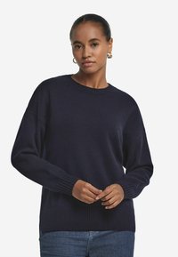 Marineblauer Strickpullover mit Rundhalsausschnitt, gerippten Bündchen und Saum. Hat eine lockere Passform und weiche Textur. Getragen mit blauen Jeans.