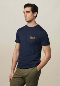 Camiseta de manga corta azul marino de tela suave con un bolsillo rectangular que presenta un logo naranja de "HACKETT". Diseño ajustado, cuello redondo.