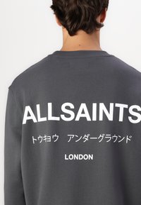 Felpa grigia con colletto a girocollo, con grande scritta bianca sul retro: "ALLSAINTS" e "LONDON", con caratteri giapponesi sottostanti.