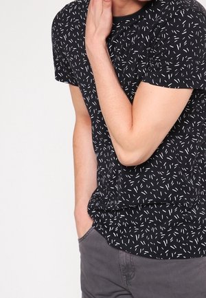 Musta t-paita, jossa on valkoinen pilkullinen kuvio, lyhyet hihat ja rento istuvuus. Yhdistetty harmaisiin housuihin.