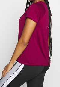 Camiseta de manga corta en color burdeos con escote redondo y un detalle de corte en la espalda, combinada con leggings negros con rayas blancas y grises.