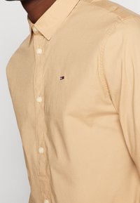 Camisa de botones beige de tela de algodón, con cuello punta y pequeño logotipo bordado en rojo, blanco y azul en el pecho.