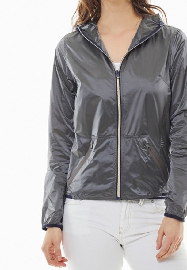 Femme portant une veste à capuche zippée gris foncé brillante avec des poches avant et un pantalon blanc, debout devant un fond blanc.