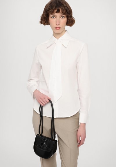 Camicia bianca a maniche lunghe con un ampio colletto, caratterizzata da un dettaglio a cravatta. Sono visibili anche pantaloni beige e una piccola borsa nera con manico.
