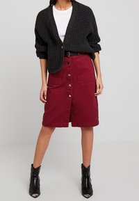 Cardigan nera a maglia strutturata sopra una maglietta bianca, abbinato a una gonna di denim bordeaux con chiusura a bottoni frontale, indossata con stivaletti neri alla caviglia.