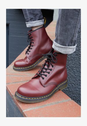 Bordeaux leren veterschoenen met een ronde neus, zwarte veters en gele stiksel, rustend op een bakstenen oppervlak.