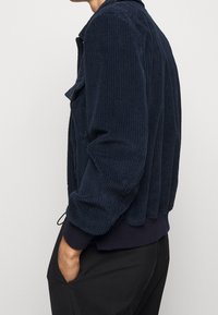 Osoba nosząca granatową kurtkę z kordurowej tkaniny, z rękami w kieszeniach czarnych spodni, showna z boku na tle jednolitym.