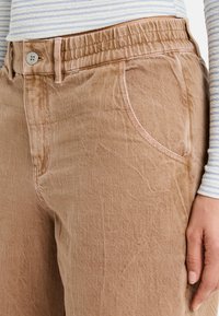 Pantalones de color marrón claro, con textura, que tienen una cinturilla elástica y bolsillos laterales. Presentan costuras en contraste y un cierre de botón en la cintura.