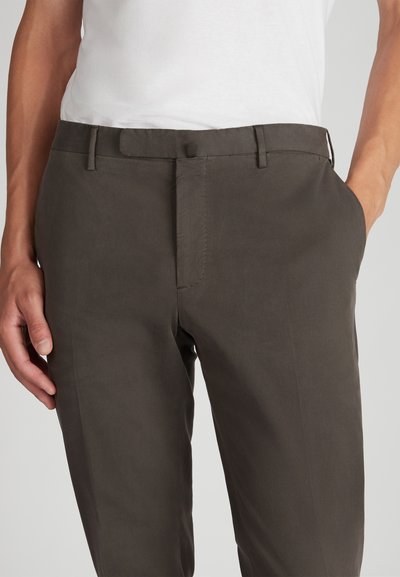 Primer plano de una persona que lleva pantalones ajustados de color marrón oscuro con bolsillos delanteros, combinados con una camisa blanca metida por dentro.