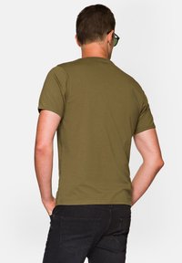 Oliwkowa koszulka z krótkim rękawem o okrągłym dekolcie, wykonana z lekkiego materiału i w luźnym kroju, noszona z ciemnymi dżinsami.