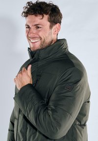 Dark green puffer jacket with a high collar, textured panels, and a chest pocket. Features a branded patch on the left shoulder.