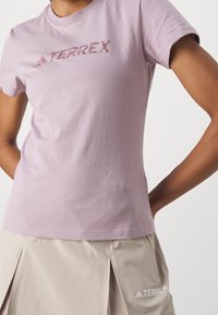 Person som bär en ljuslila TERREX-t-shirt och en beige kjol med TERREX-logotyp, med händerna placerade bakom ryggen, stående mot en vit bakgrund.