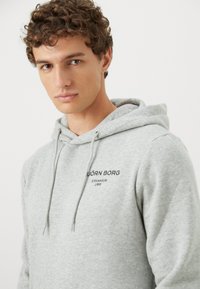 Młody mężczyzna z kręconymi włosami, ubrany w jasnoszary hoodie z napisem "BJÖRN BORG STOCKHOLM 1956" na piersi.