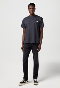 Vyriški pilki marškinėliai trumpomis rankovėmis su apvalia apykakle, puošiami baltu „Wrangler“ logotipu, derinami su juodais siaurais džinsais ir juodais sportbačiais.