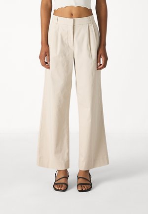 Pantalons larges beige à taille haute et plis, fabriqués en tissu léger. Assortis à des sandales noires à lanières.