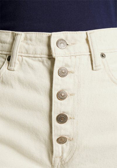 Zbliżenie beżowych dżinsów z pięcioma metalowymi guzikami i pętelkami na pasek, noszonych z ciemnogranatową bluzką.