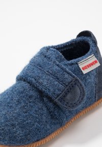 Chausson en feutre bleu avec sangle velcro. Il présente une surface texturée, une semelle en caoutchouc et un accent en tissu gris. Étiquette de marque sur le côté.