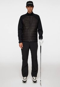 Svart och mörkblå zip-up jacka med vadderad framdel och långa ärmar. Svarta byxor, vita och svarta sneakers, golfklubba i höger hand, vit handske.