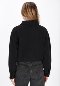Černá, oversized košile s límcem, vyrobená z měkké tkaniny. Má spuštěná ramena a zadní část s yoke detailem, zkombinováno s tmavě šedými džíny.