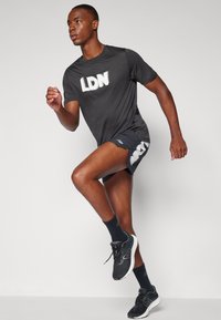 Tamsiai pilkas sportinis marškinėliai su "LDN" užrašu, derinami su juodomis šortais, turinčiais baltą akcentą. Juodi sportbačiai ir kojinės užbaigia aprangą.