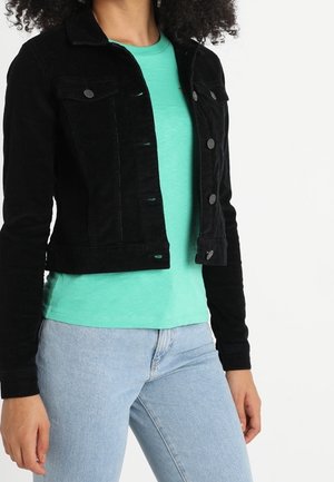 Vrouw die een zwarte jasknop draagt over een turquoise t-shirt en lichtblauwe spijkerbroek, staand tegen een witte achtergrond.