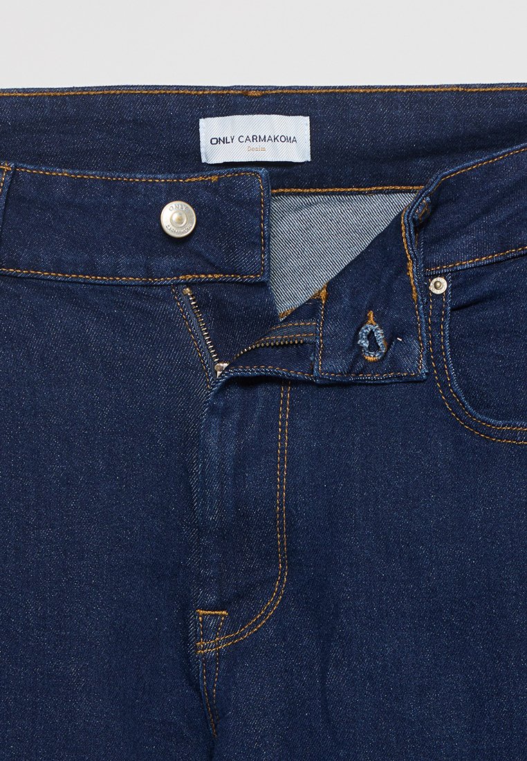 Jeans en denim bleu foncé avec une fermeture éclair, des coutures en cuivre et une étiquette de marque au niveau de la taille. Design classique à cinq poches.