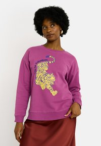 Mujer con cabello rizado vestida con un sudadera púrpura con un tigre amarillo y el texto "La vida es libertad", combinada con una falda roja, posando frente a un fondo blanco.