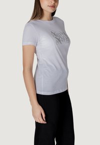 T-shirt bianca in cotone con maniche corte, caratterizzata da un logo argentato lucido sul petto, scollo rotondo e silhouette aderente.