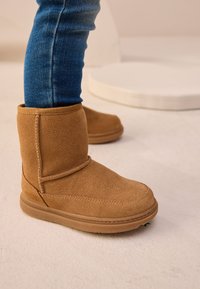 Ghete scurte din piele întoarsă de culoarea bronzului, cu vârf rotund, accente cusute și talpă de cauciuc, asortate cu blugi albaștri skinny. Suprafață texturată vizibilă.