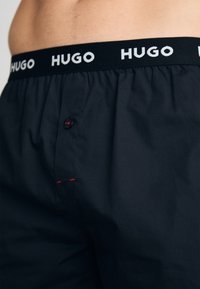 Boxeri negri cu un elastic alb inscripționat "HUGO", având închidere cu nasture și o cusătură roșie accentuând zona de lângă nasture. Textură fină.