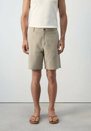 Hombre con pantalones cortos beige hasta la rodilla, camiseta blanca sin mangas y chanclas marrones, de pie frente a un fondo liso.