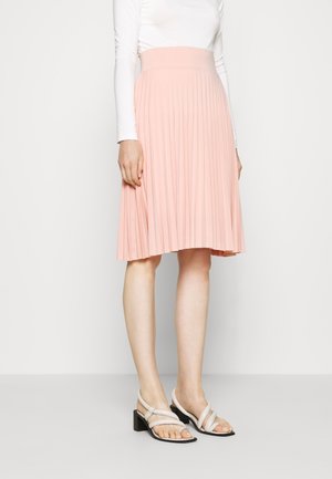 Lichtroze geplooide rok met een hoge tailleband, gemaakt van zachte stof. Paar witte sandalen met bandjes en een lage blokhak.