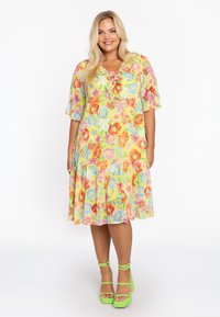 Blumenmusterkleid in lebhaften Farben, mit kurzen Ärmeln, einem Rüschensaum und einem V-Ausschnitt mit Bindeband. Kombiniert mit grünen Riemchensandalen.