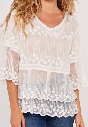 Blouse blanche transparente avec broderie florale, bords festonnés et manches volantées, portée sur un débardeur et un jean bleu.