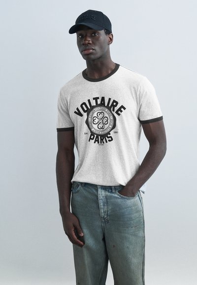 Νέος άντρας που φοράει λευκό T-shirt με περιθώρια και το κείμενο «Voltaire Paris» με έμβλημα, συνδυασμένο με μπλε τζιν και μαύρο καπέλο baseball.