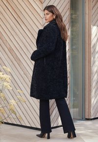 Palton negru din blană sintetică cu un finisaj texturat, guler și buzunare laterale, asortat cu pantaloni închiși la culoare și pantofi cu toc înalt, în fața unui fundal din panouri de lemn.