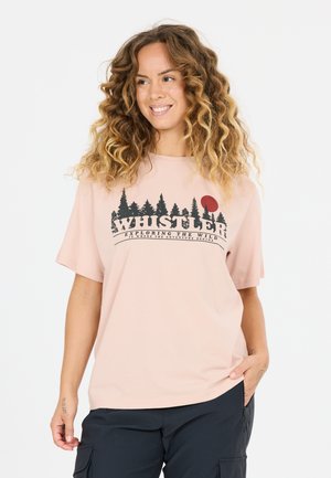 Smilende kvinde med krøllet hår iført en lyserød Whistler T-shirt med træer og en rød sol, poserer med den ene hånd i lommen.