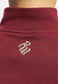 Bordo megztinis su aukštu apykakle ir švelniu tekstūra, su smėlio spalvos logotipu išsiuvinėtame gale netoli kaklo.