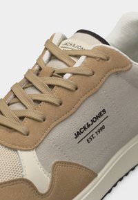 Εγγύς πλάνο μπεζ και καφέ αθλητικού παπουτσιού Jack & Jones με υφασμάτινες και σουέντ υφές, δεμένα κορδόνια και λογότυπο στην πλευρά.