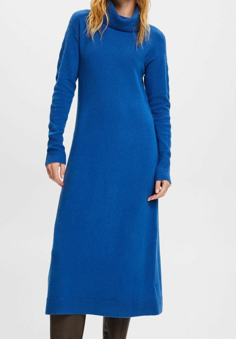 Femme portant une robe-pull longue en maille bleue avec un col roulé et des manches longues, accompagnée de leggings ou pantalons foncés.