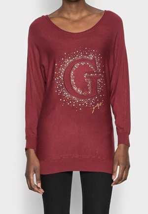 Pull à manches longues rouge foncé avec une grande lettre G ornée et un petit texte doré sur le devant, porté par une personne avec un pantalon noir.